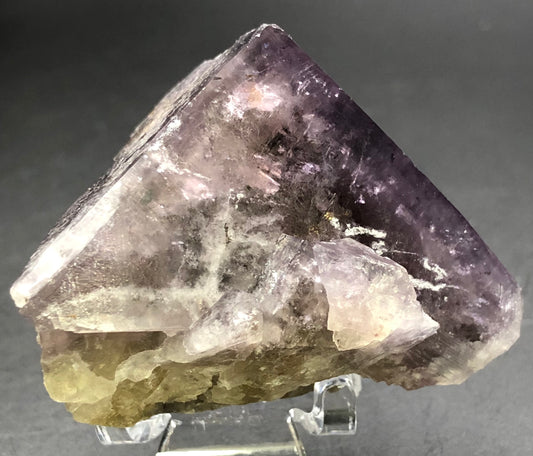 A large, rough fluorite gemstone from The Crystalary's AUCTION collection sits on a clear display stand against a dark background. With shades of purple, white, and brown, its triangular form reveals fascinating translucency and mixed smooth and jagged surfaces.