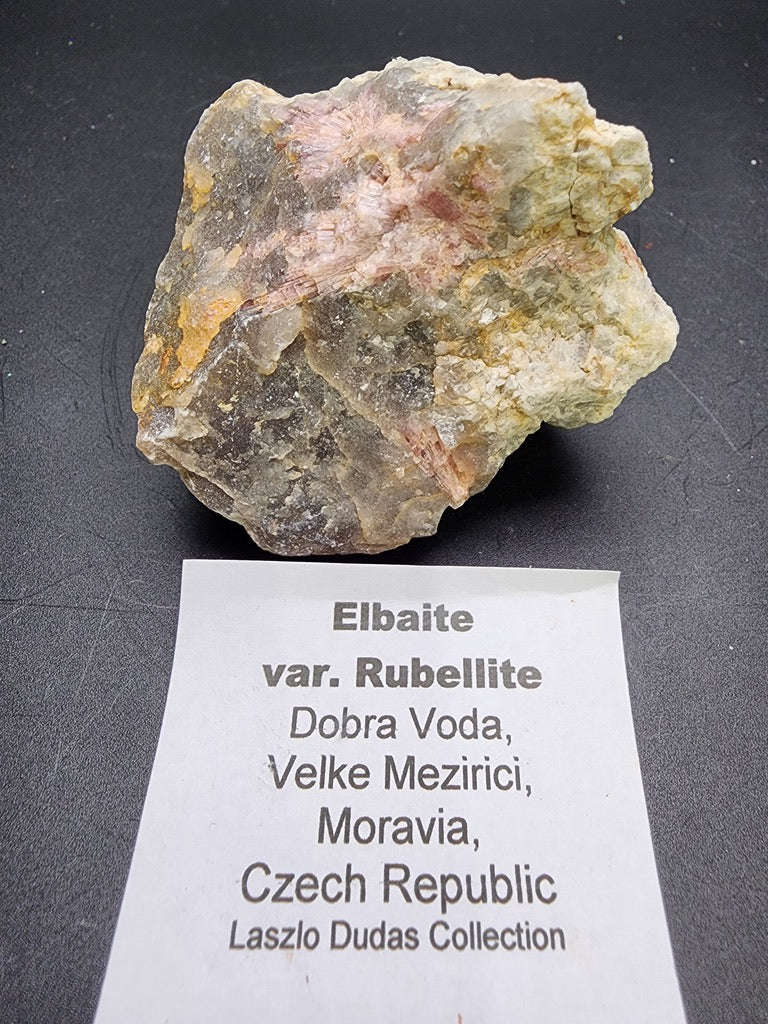 On display is a rough stone specimen from The Crystalary, labeled "Elbaite var. Rubellite," featuring delightful shades of pink and white with its natural texture preserved. Originating from Dobra Voda, Velke Mezirici, Moravia, Czech Republic, this piece is an esteemed item in the Laszlo Dudas Collection.