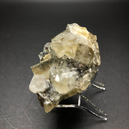 A stunning display from The Crystalary showcases a fluorite cluster from The Bull Vein at the Lady Annabella Mine in Eastgate, Weardale, Co. Durham, England. This remarkable piece features translucent crystals with angular formations elegantly set on a clear stand against a dark backdrop. Predominantly colorless, the crystals exhibit subtle yellowish and gray hues that mesmerize under changing daylight conditions.