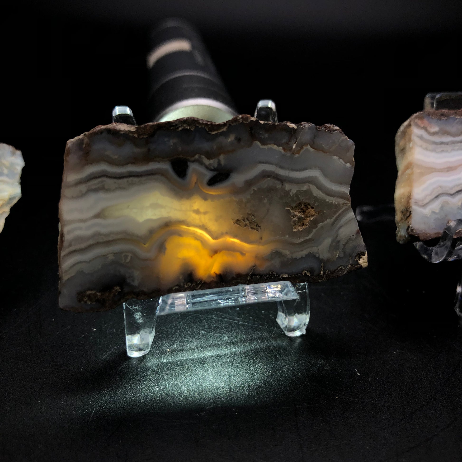 A slice of the Fourth of July Peak agate from Hassayampa, Maricopa County, Arizona—part of The Crystalary’s collection—is backlit by a light source. This reveals its intricate patterns and warm, glowing colors. Displayed on a clear stand against a dark background, this stunning piece from The Crystalary is sure to captivate any viewer.