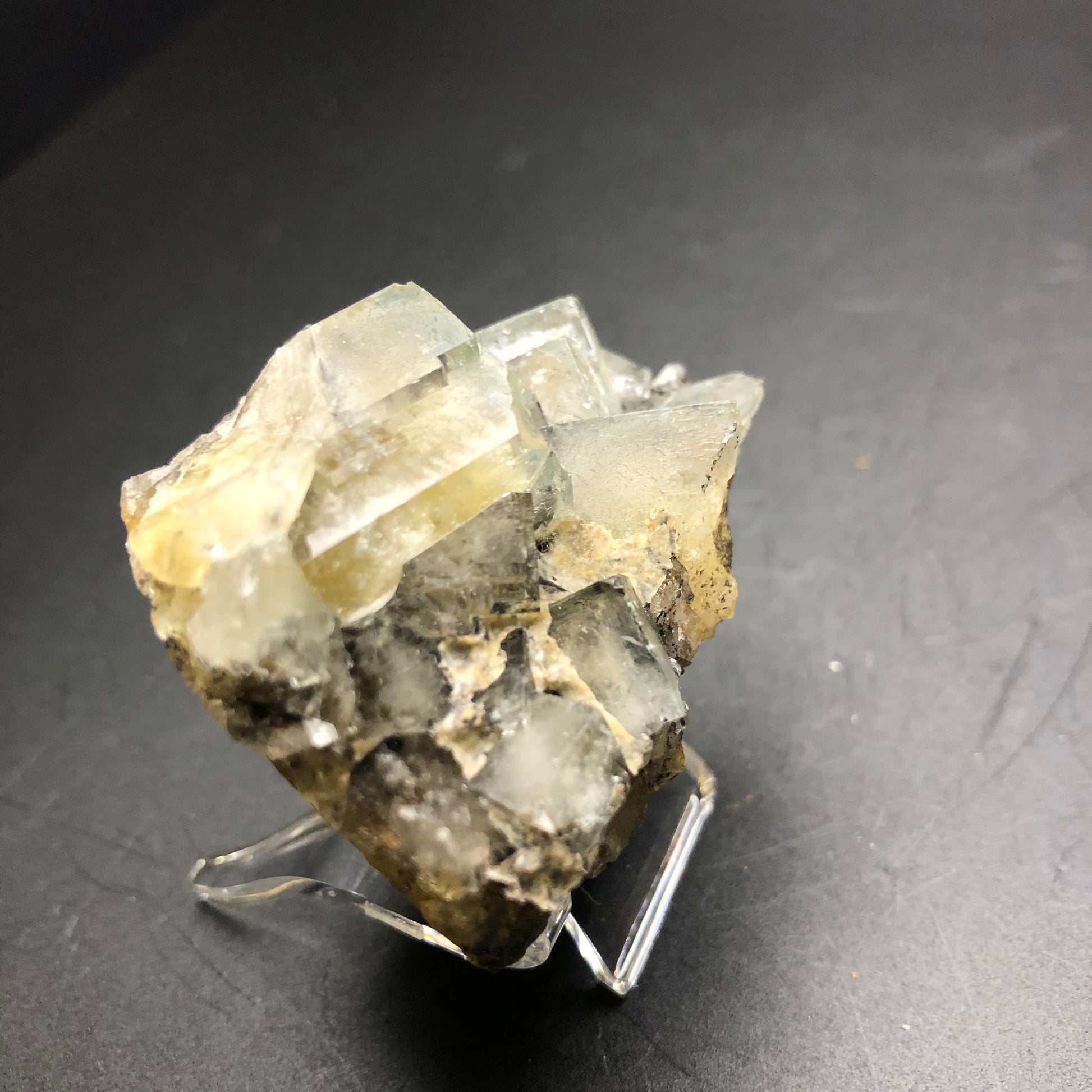 A cluster of translucent fluorite crystals, sourced from The Bull Vein at the Lady Annabella Mine in Eastgate, Weardale, Co. Durham, England, rests on a clear stand against a dark background. Sold by The Crystalary, these crystals have rough edges and showcase varying sizes with a pale yellow hue accented by greenish tones.