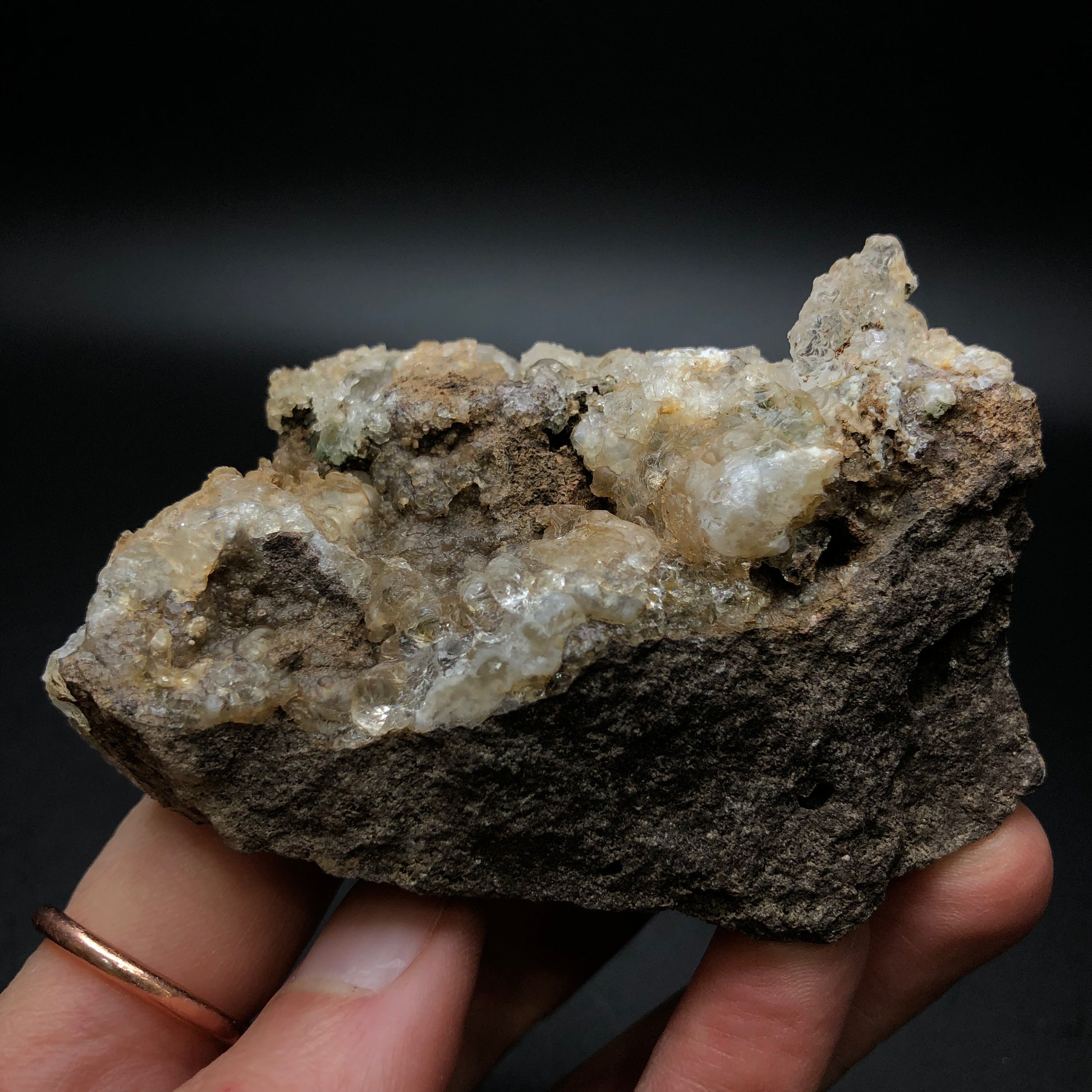 In a hand, an unrefined rock featuring white, translucent crystals is held against a dark backdrop. These fragile structures, identified as stalactic hyalite opal from La Ventilla in the Villa de Reyes Municipality of San Luis Potosí, Mexico, showcase an enchanting green fluorescence that captures the mineral splendor of the region. This exquisite piece is offered by The Crystalary under the name AUCTION- Hyalite.