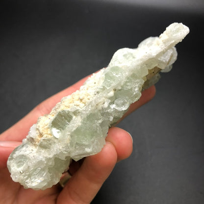 A hand holds a translucent, greenish-white crystal cluster from The Crystalary's AUCTION- Fluorite collection, highlighting its rough texture and geometric formations against a dark background. One side of the crystal features an earthy tone, similar to the micro-quartz matrix structures characteristic of those discovered in the renowned Xianghualing Mine in Linwu Co., Chenzhou, Hunan, China.