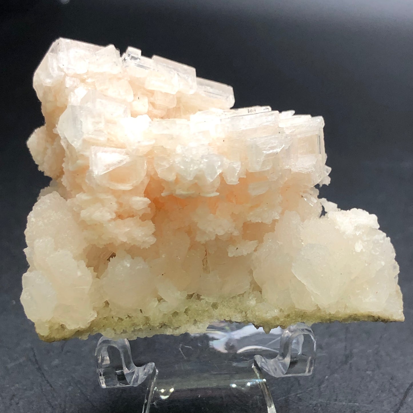 A close-up of the "AUCTION- Halite- unk locality" from The Crystalary reveals a pale pink and white mineral formation with crystalline structures. It is elegantly displayed on a clear stand against a dark background, showcasing halite crystals in an isometric formation with a rough, jagged appearance.