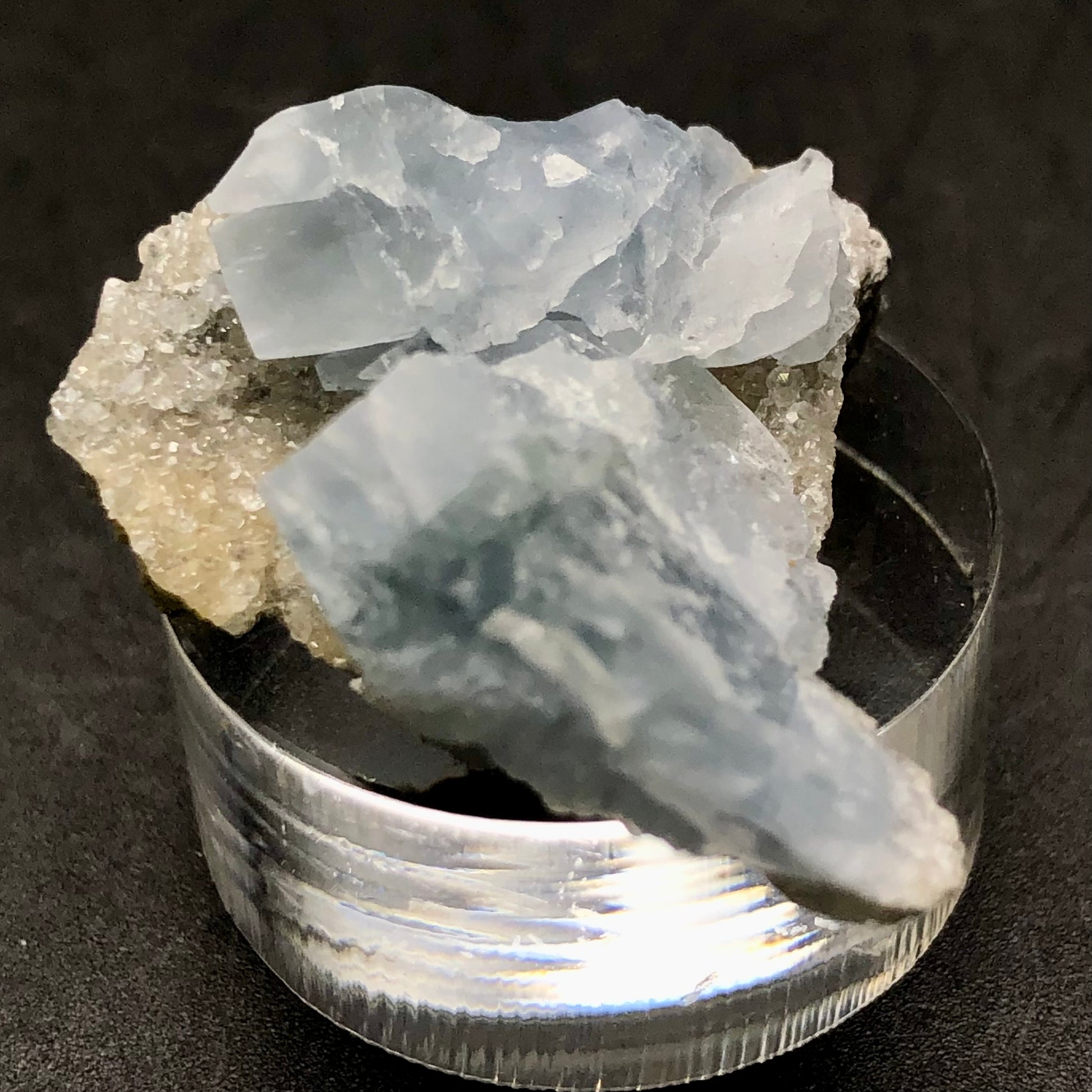 A cluster of pale blue Celestine crystals from The Crystalary is displayed on a transparent cylindrical stand against a black background, bearing a rough texture and slightly translucent appearance that captures the allure similar to treasures found in Meckley's Quarry.