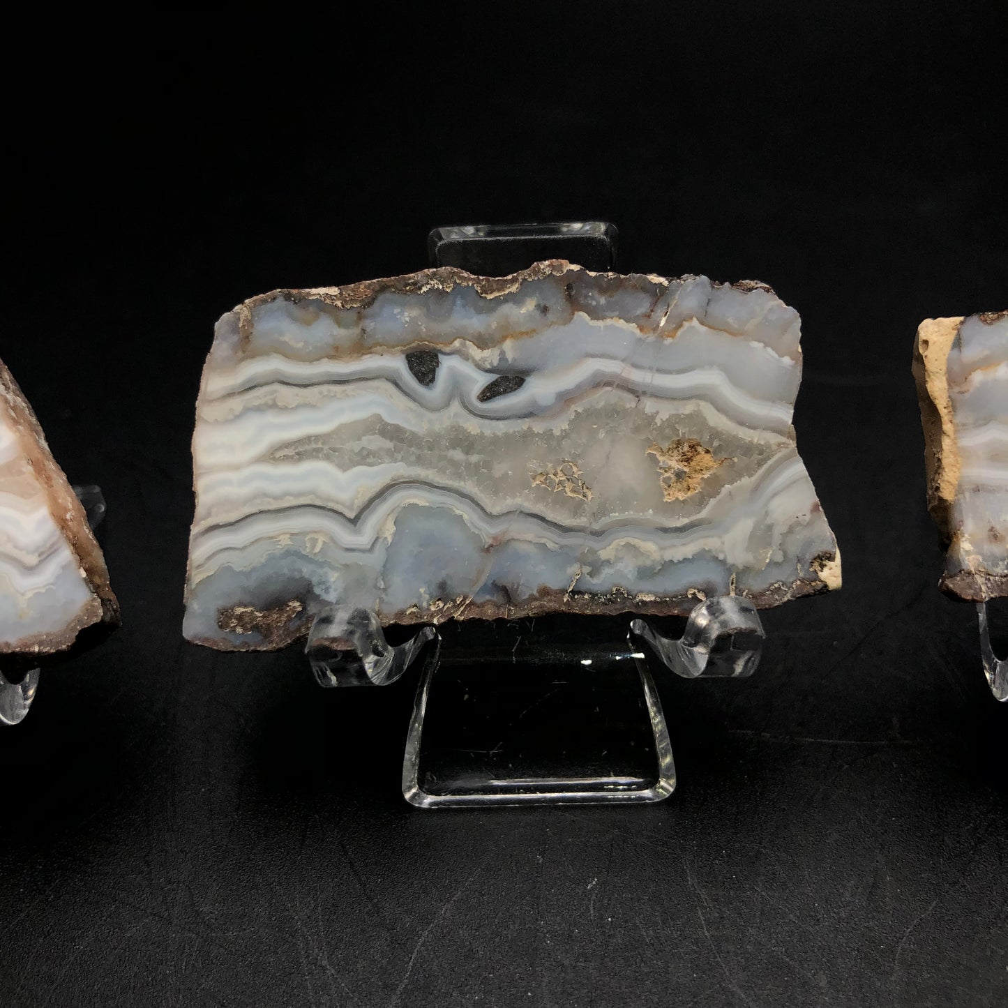 A refined slab of agate from the Fourth of July Peak collection, featured in The Crystalary auction, showcases intricate gray, cream, and beige patterns as it rests on a clear stand against a black backdrop.