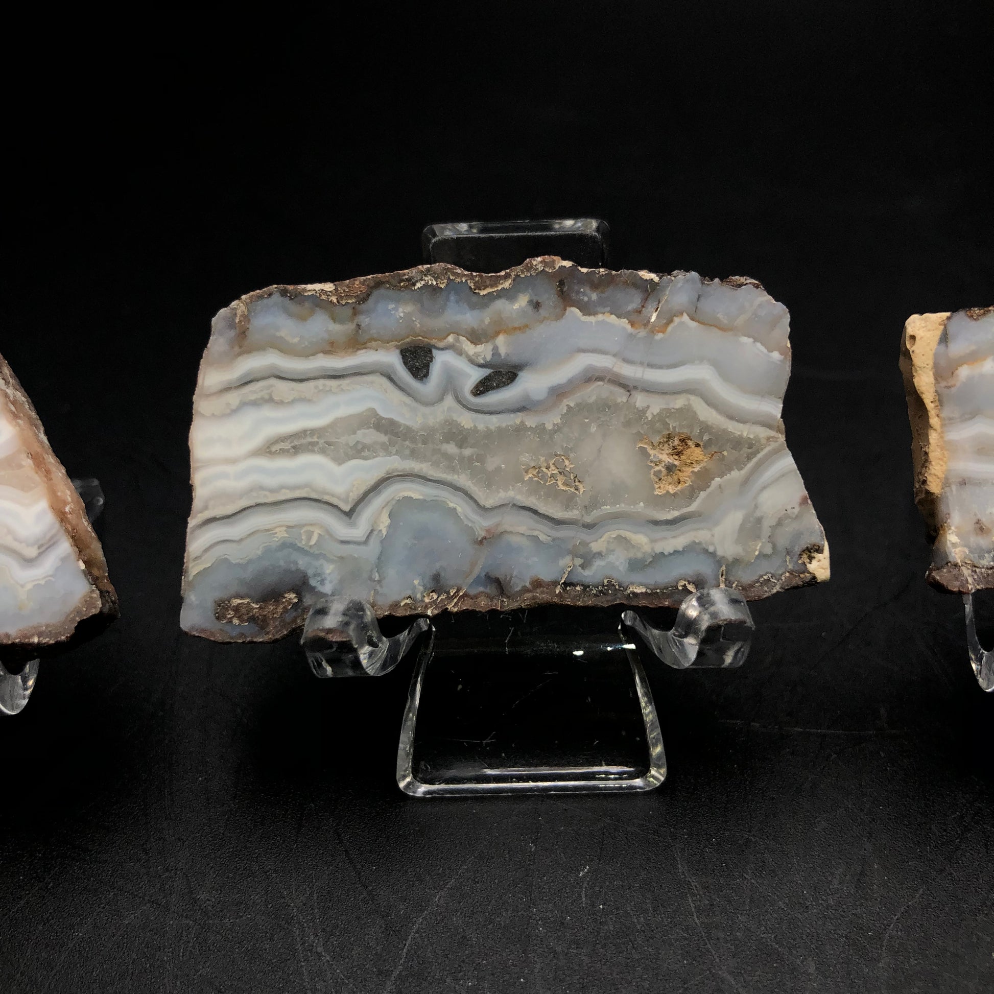 A refined slab of agate from the Fourth of July Peak collection, featured in The Crystalary auction, showcases intricate gray, cream, and beige patterns as it rests on a clear stand against a black backdrop.