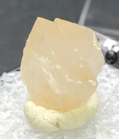 A close-up shows a rough, translucent calcite crystal from Otero County, Colorado, USA by The Crystalary. It features a pale peach color with a light honey hue on a beige clay-like base and textured white surface against a dark background.