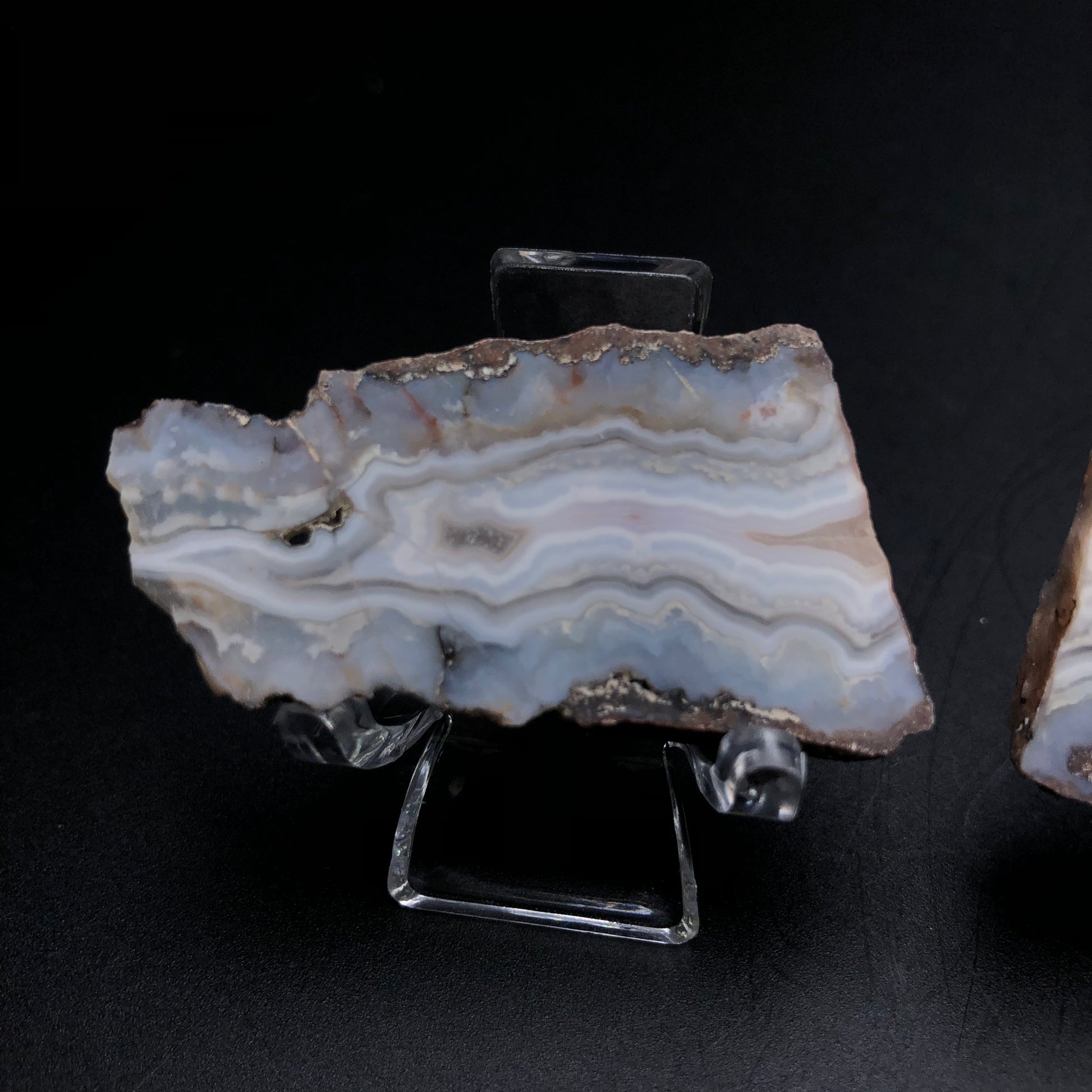 A slice of agate from Fourth of July Peak, beautifully showcased on a clear stand against a black backdrop. This piece from The Crystalary features layered patterns in shades of blue, white, and gray with intricate banding and a rough edge that captures the natural beauty of Fourth of July Butte in Arizona.