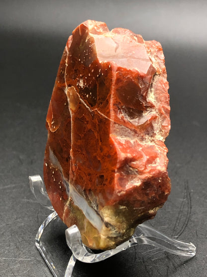 A stunning product from The Crystalary, named "AUCTION- Agate- unk locality," features a polished red agate slice with reddish-brown hues and white and beige streaks, elegantly displayed on a clear stand against a dark background.