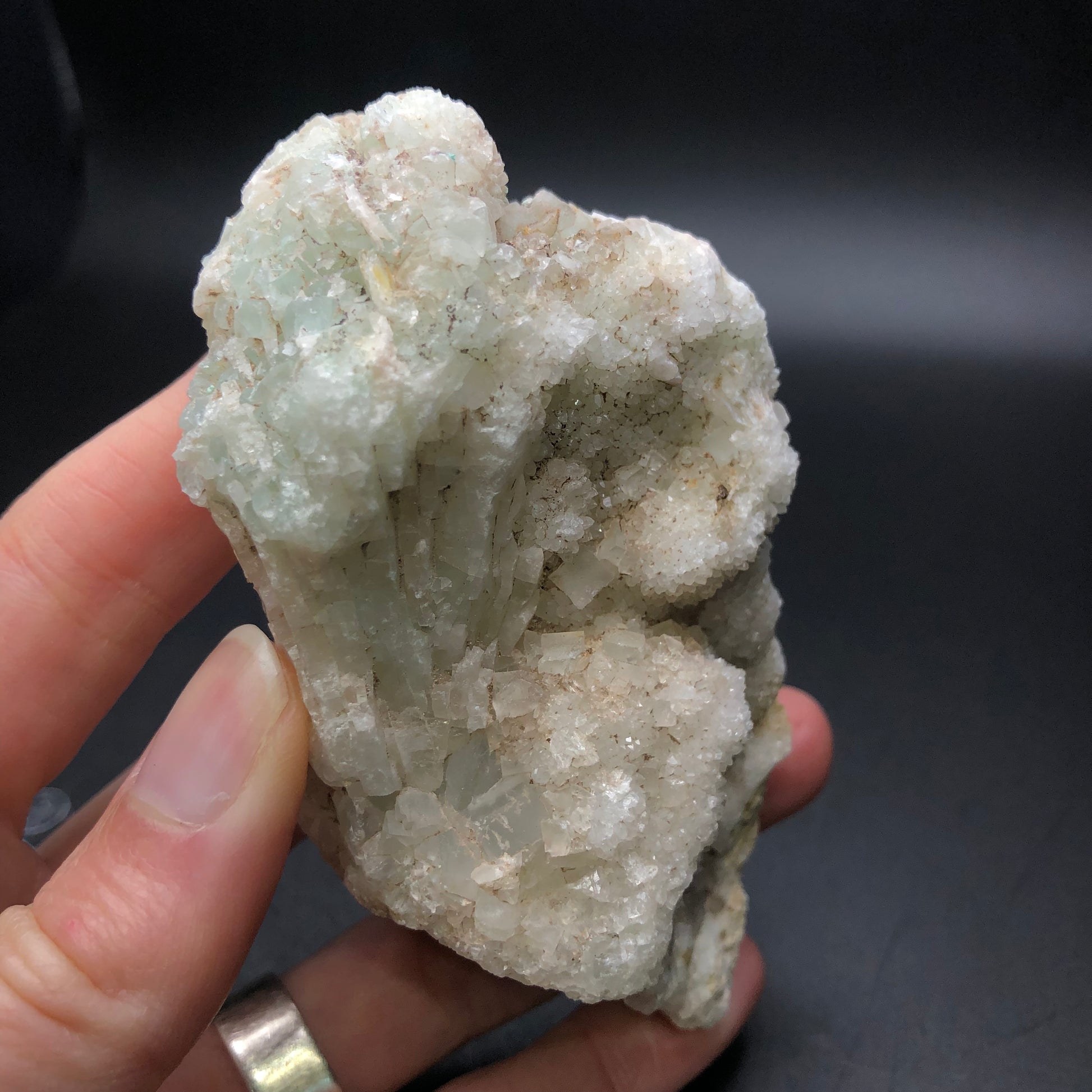 A hand holding a mineral specimen from The Crystalary, featuring an AUCTION- Calcite and Quartz with a light greenish hue and crystalline texture, reminiscent of druzy white quartz, against a dark background.