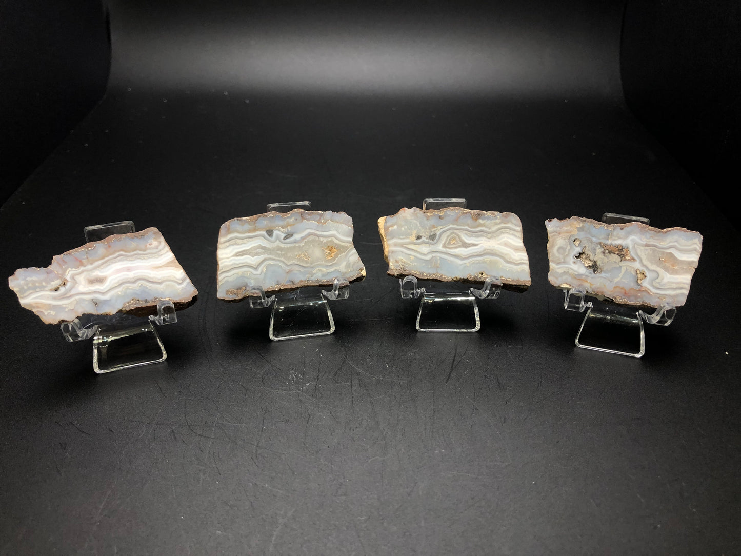 Four slices of AUCTION- Agate from Fourth of July Peak, showcased on display stands against a dark backdrop. These slabs from The Crystalary exhibit intricate layered patterns in gray and white, reminiscent of the breathtaking vistas at Fourth of July Peak in Hassayampa, Maricopa County, Arizona.