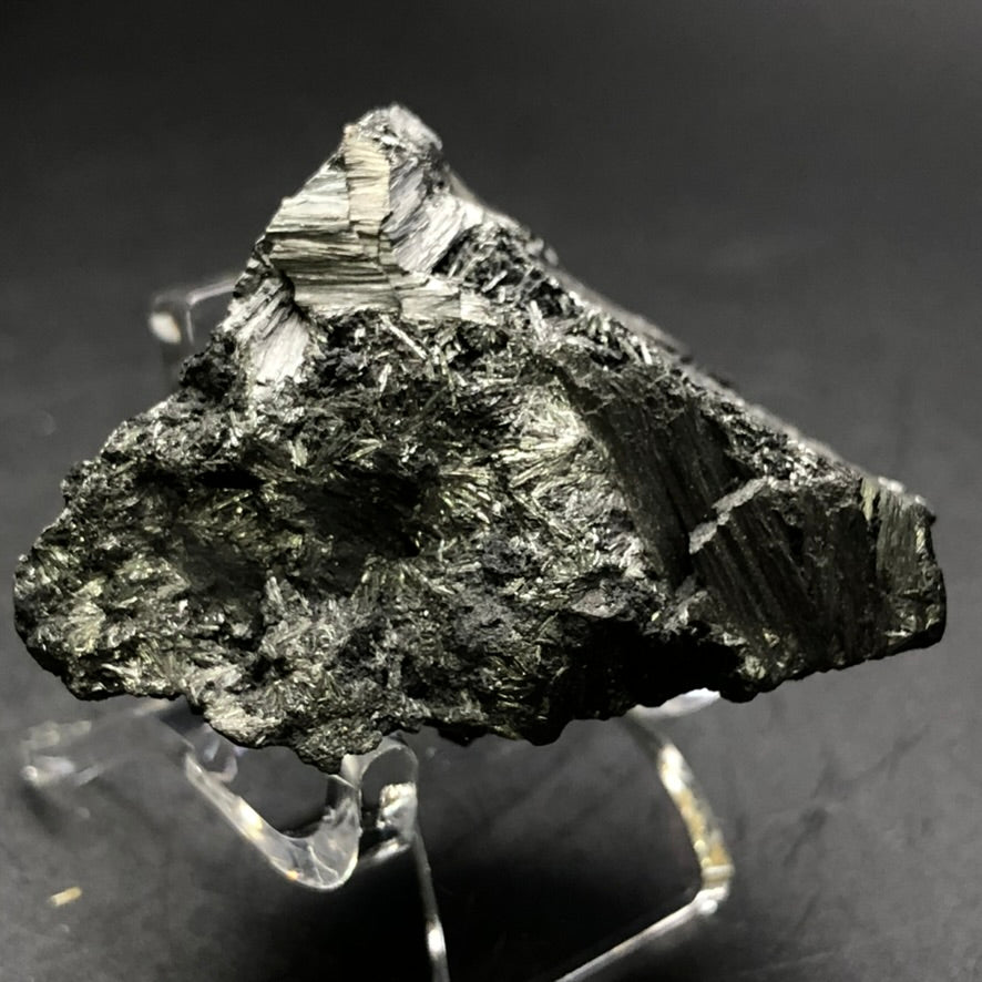 A detailed photograph showcases the AUCTION- Pyrolusite from Elgersburg, Ilm District, Thuringia, Germany by The Crystalary. The black mineral with its rough texture is mounted on a clear stand against a dark background. Its surface displays angular and layered designs, featuring radiating crystals that enhance its complex appearance.