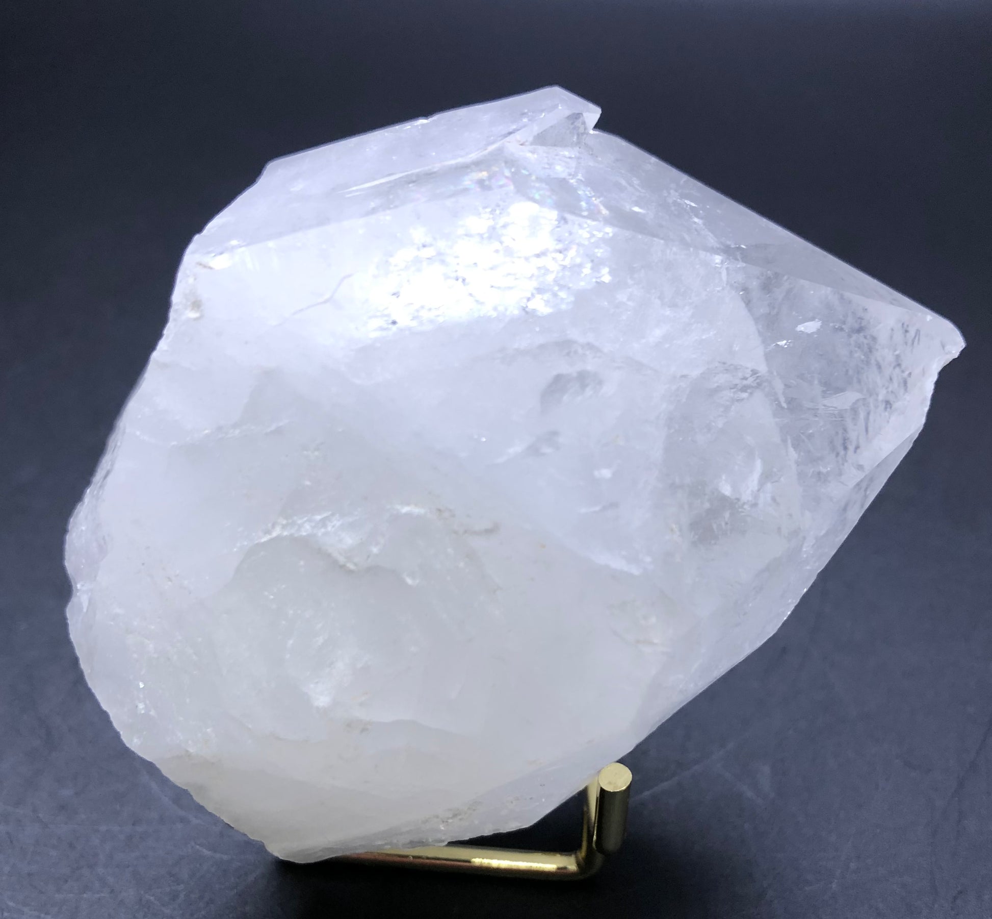 A large, translucent white quartz point from The Crystalary is shown against a dark background with rough, jagged surfaces and slightly reflective facets on a small golden stand. Known as AUCTION- Quartz- unk locality.