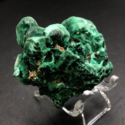 A striking malachite specimen from The Crystalary, sourced from the Liufengshan Mine in the Guichi District of Chizhou, Anhui, China, showcases its vibrant green hues and swirling patterns. Set on a clear stand against a dark backdrop, this natural stone displays a rugged texture and various shades of green.