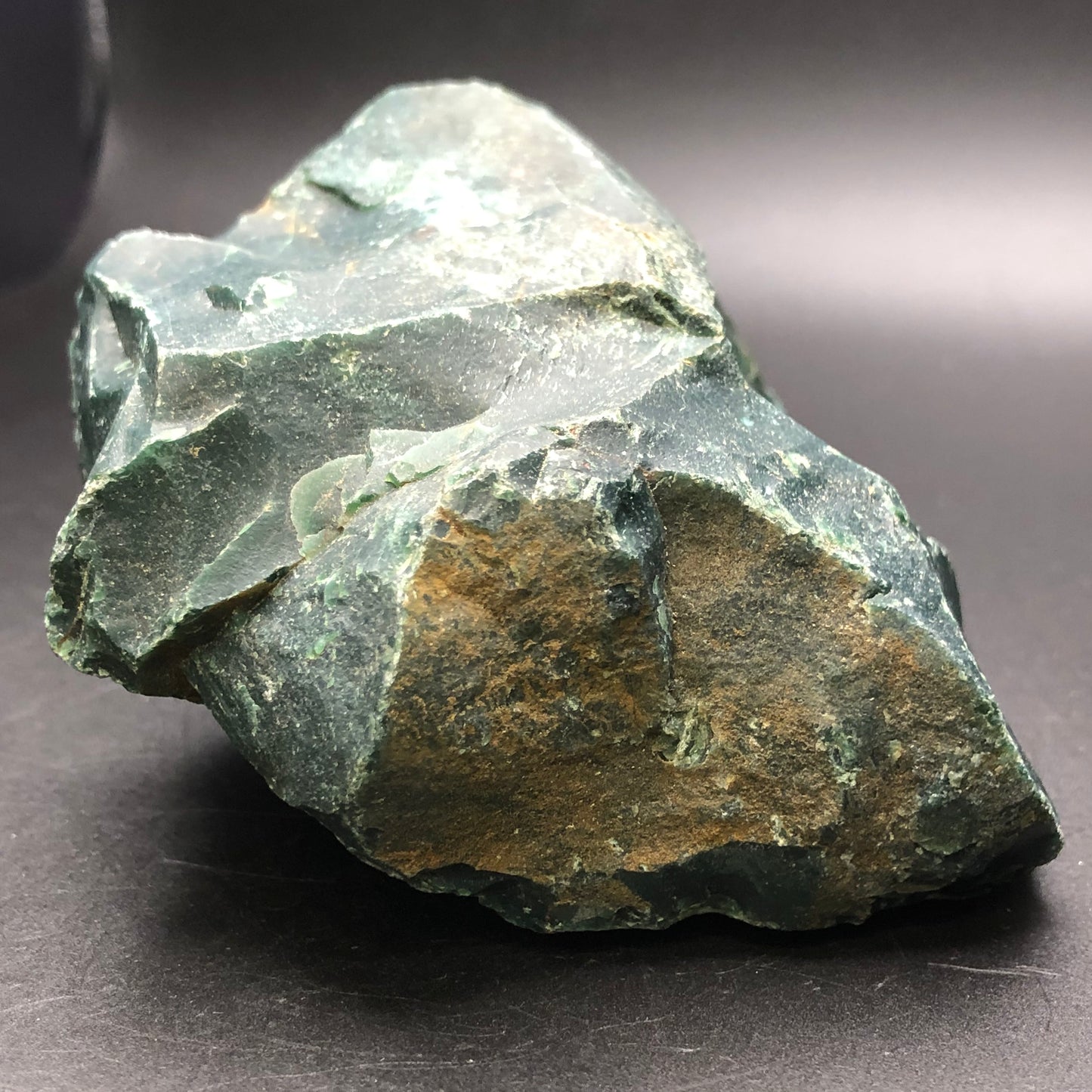 A rough, unpolished specimen from The Crystalary, the AUCTION- Bloodstone- unk locality, with brown patches set against a dark background, showcases its jagged edges and natural texture.