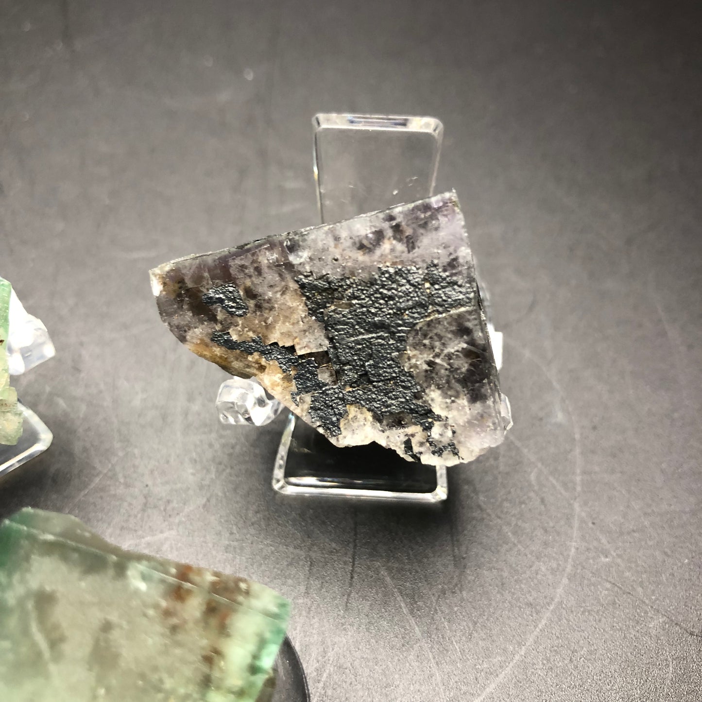 Displayed on a clear stand against a dark background, The Crystalary presents an AUCTION item – a rough, triangular fragment of fluorite from the Lady Anabella Mine in Eastgate Quarry, Eastgate, Stanhope, County Durham, England. Its mix of gray and black textures is complemented by partially visible cubes of other rocks nearby, adding to the rugged beauty of this County Durham treasure.