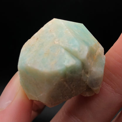 A person delicately grips a small, rough Amazonite specimen with a vitreous luster from The Crystalary between their fingers. The light blue-green mineral from Lake George, Park County, Colorado, features a few natural imperfections that beautifully contrast against the black background.