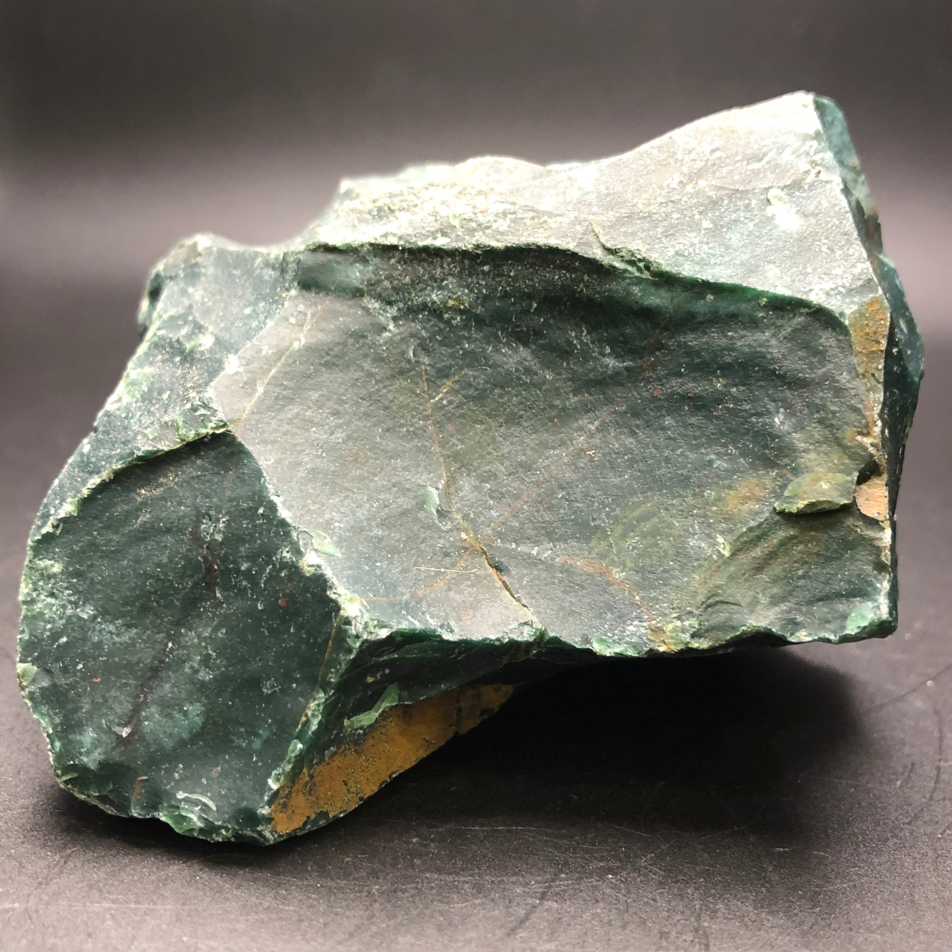 A large, jagged **AUCTION- Bloodstone- unk locality** from **The Crystalary** exhibits deep green hues and natural cracks against a dark backdrop. Lighter and darker tones intermingle across its rough surface, with brown patches that accentuate its raw beauty.