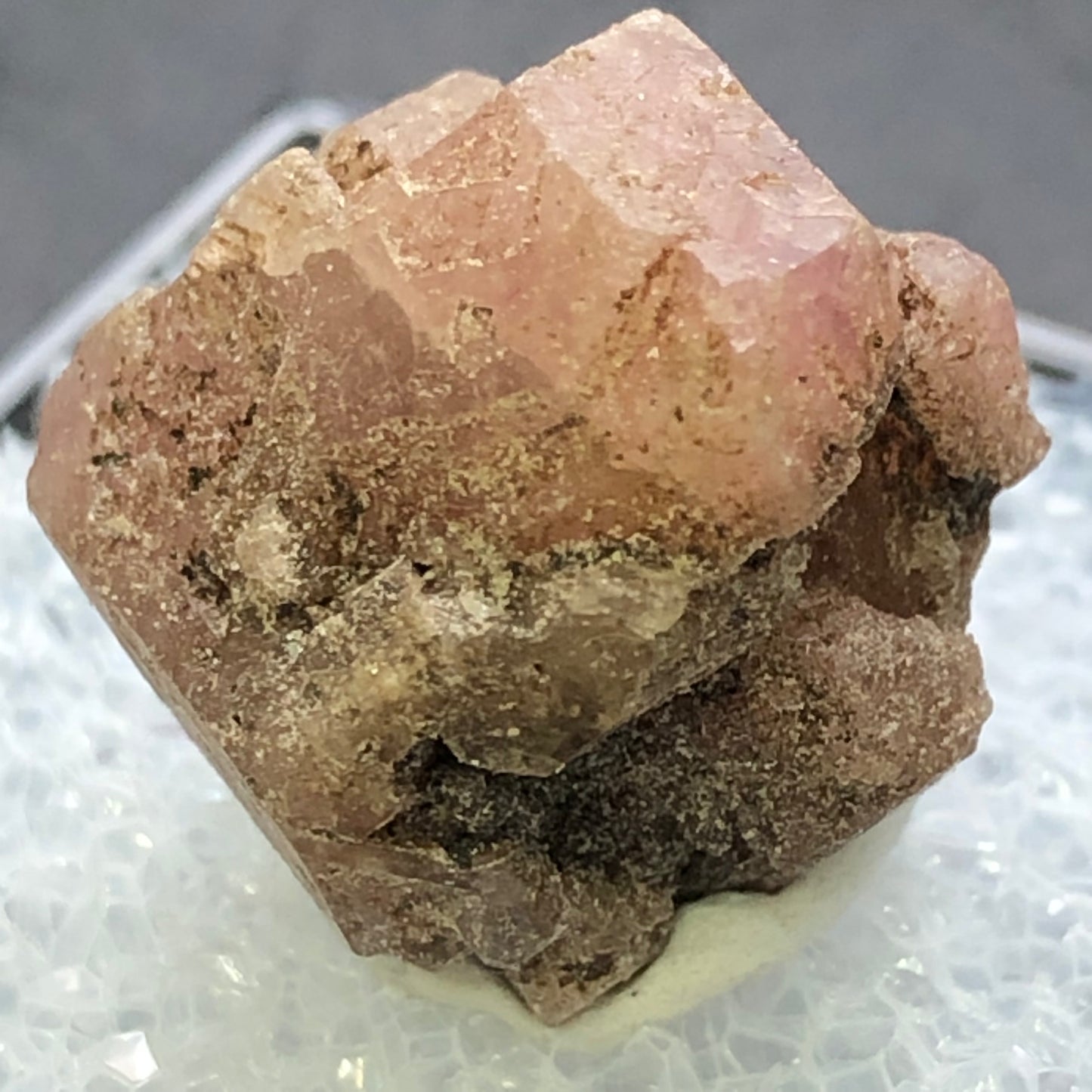 A rough garnet crystal with a dusty rose hue rests on a textured white surface, featuring an irregular shape with multiple facets and a sparkle. Set against a blurred gray background, this piece resembles garnets from Morelos, Mexico. Product: AUCTION- Garnet, Grossular by The Crystalary.