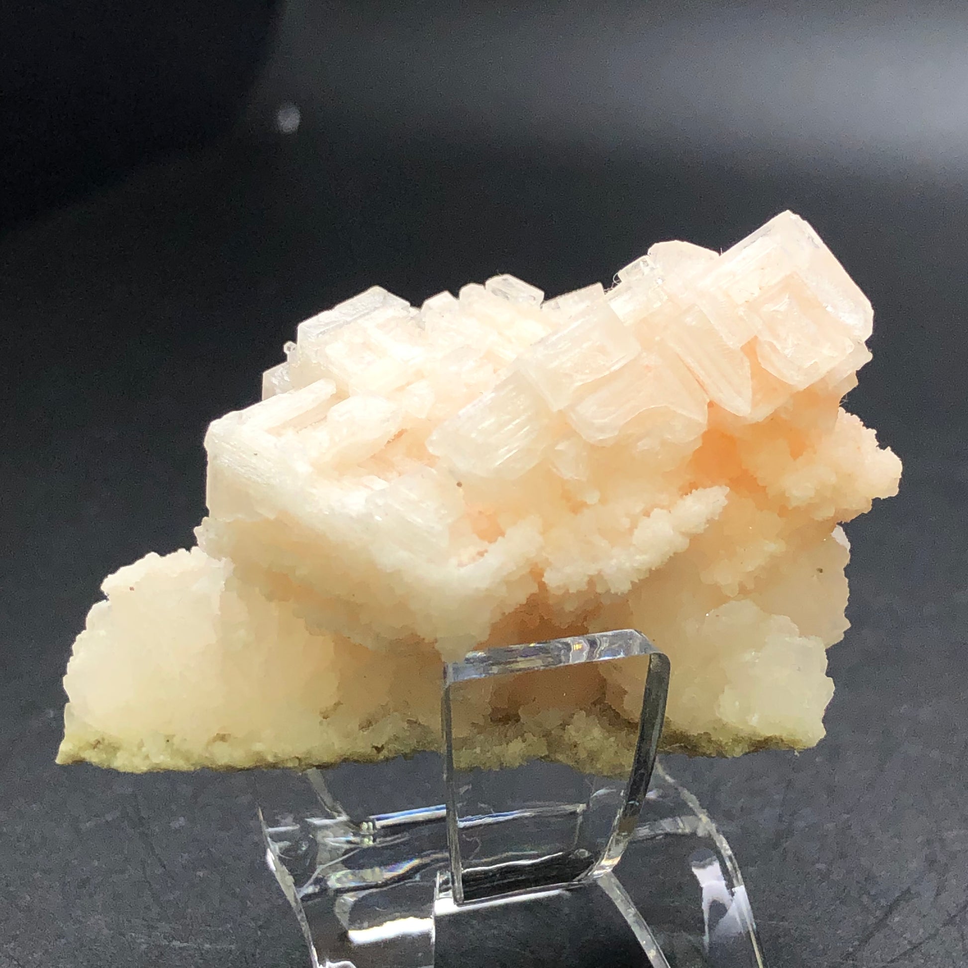 A detailed view of a translucent, pale peach-colored halite crystal from The Crystalary reveals its blocky, isometric structures set against a black background, beautifully mounted on a clear stand. The surface of this mineral appears textured and intricate.