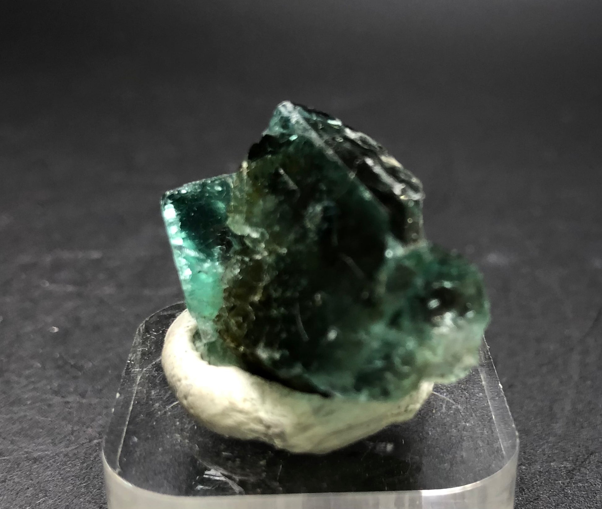 A close-up of the rough, dark green fluorite gemstone from The Crystalary reveals jagged edges as it sits on a small white base against a dark background. This piece, originating from the Diana Maria Mine in Frosterley, Weardale, County Durham, England, showcases its translucent nature and subtle daylight color-changing effects on its reflective surfaces.