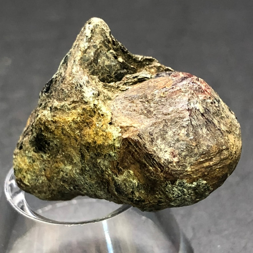 An unpolished Almadine Garnet mineral from Markstay, Ontario by The Crystalary, with a metallic sheen and mix of brown, yellow, and gray hues sits on a clear stand against a black background. Its rugged surface appears uneven.