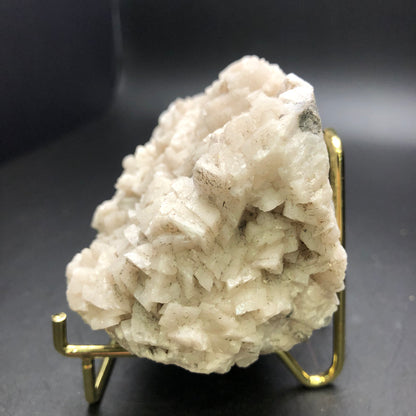 A detailed view of the AUCTION- Dolomite & Calcite from Thunder Bay, Ontario, Canada, by The Crystalary showcases a large white calcite cluster with layered formations on a gold-colored stand against a dark background, where the textured surfaces reflect light subtly.