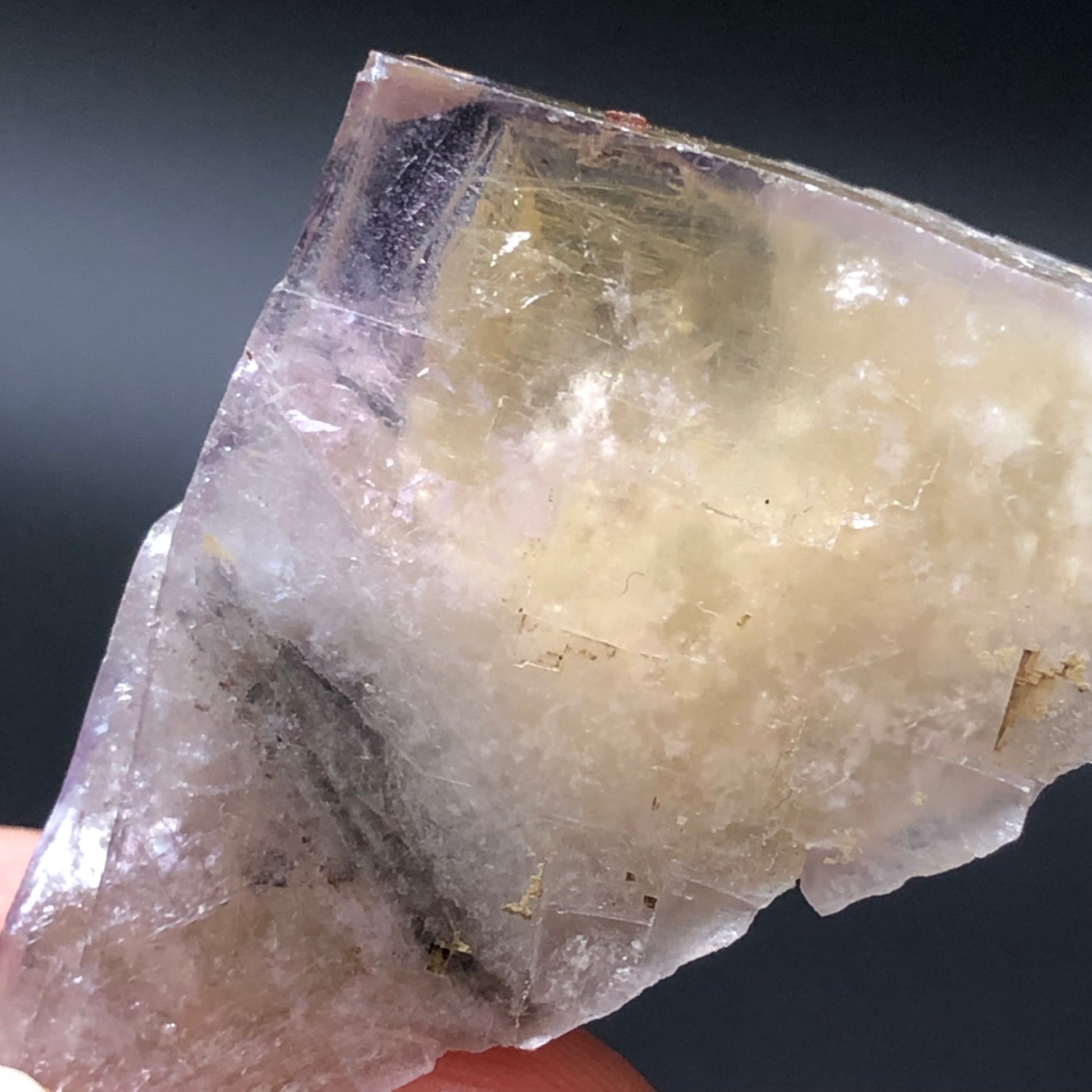 The Crystalary presents a close-up of the Fluorite from Cromwell Pocket at Greenlaws Mine, showcasing its jagged crystal form with purple exterior zoning and a golden interior. The clear, pale yellow, and purple hues highlight its natural fractures and layers against a dark backdrop.