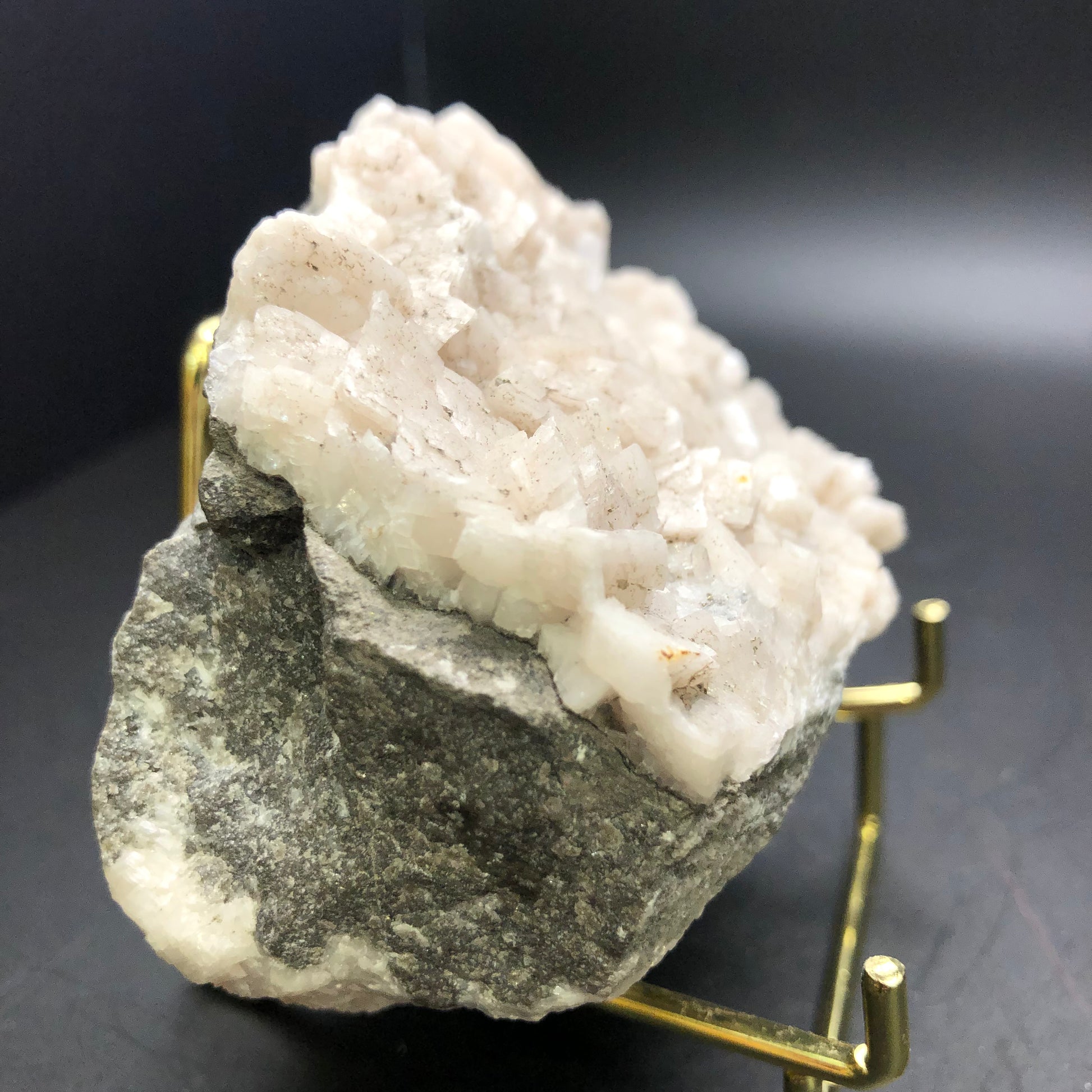 On a gold stand rests a substantial mineral specimen from The Crystalary, featuring Dolomite and Calcite crystals. The piece displays a rough gray base adorned with an upper layer of white crystalline formations. Its backdrop captures the tranquil and neutral tones reminiscent of Thunder Bay's landscapes in Ontario, Canada.