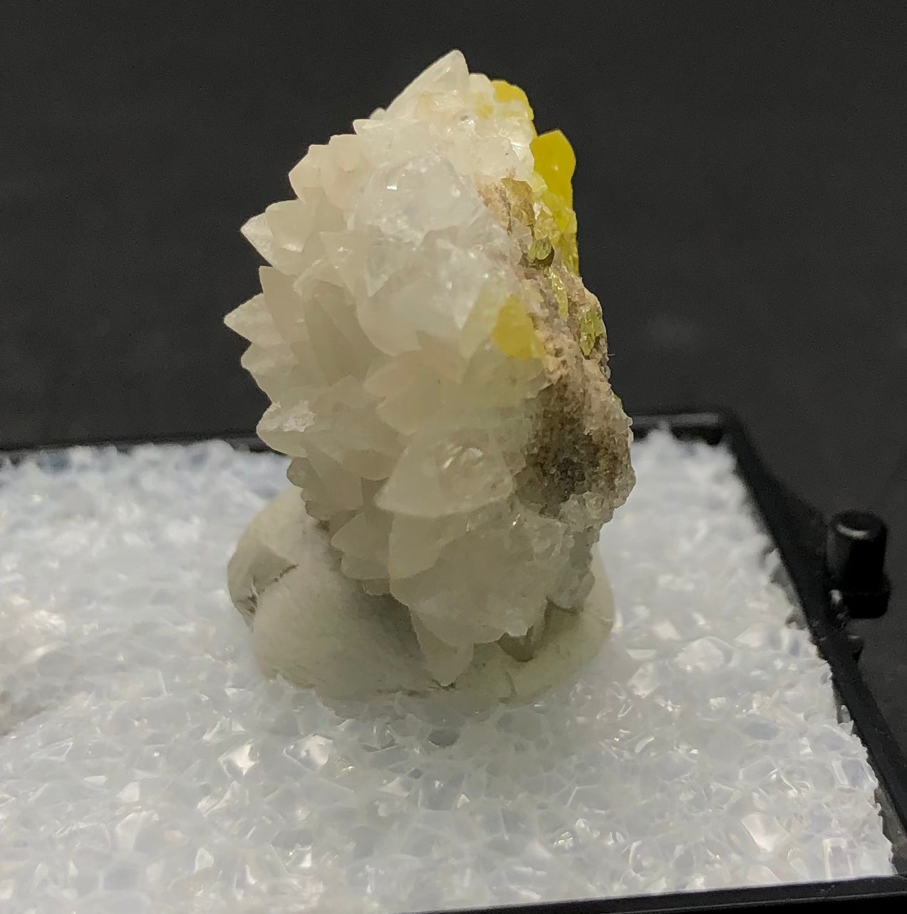 A close-up captures a small cluster of translucent, pointed calcite crystals with a hint of yellow, suggestive of sulfur. This stunning specimen from Maybee Quarry in Exeter Township, Michigan is elegantly presented on a granular white surface within a display case, courtesy of The Crystalary.