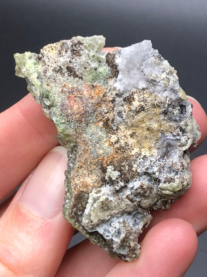 A close-up of a hand holding a specimen of the AUCTION- Smithsonite var. Cuprian from the 79 Mine in Chilito, located in the Hayden area, Banner Mining District, Gila County, Arizona. This rough mineral rock by The Crystalary showcases a textured surface with earthy shades of grey, brown, and green.