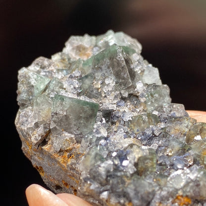 A close-up image of a person holding The Crystalary’s Fluorite from the Northern Lights Pocket at Diana Maria Mine, Co. Durham, England. The mineral cluster showcases cubic and crystalline formations with crystals in greenish and clear hues that brilliantly reflect light against a blurred background.