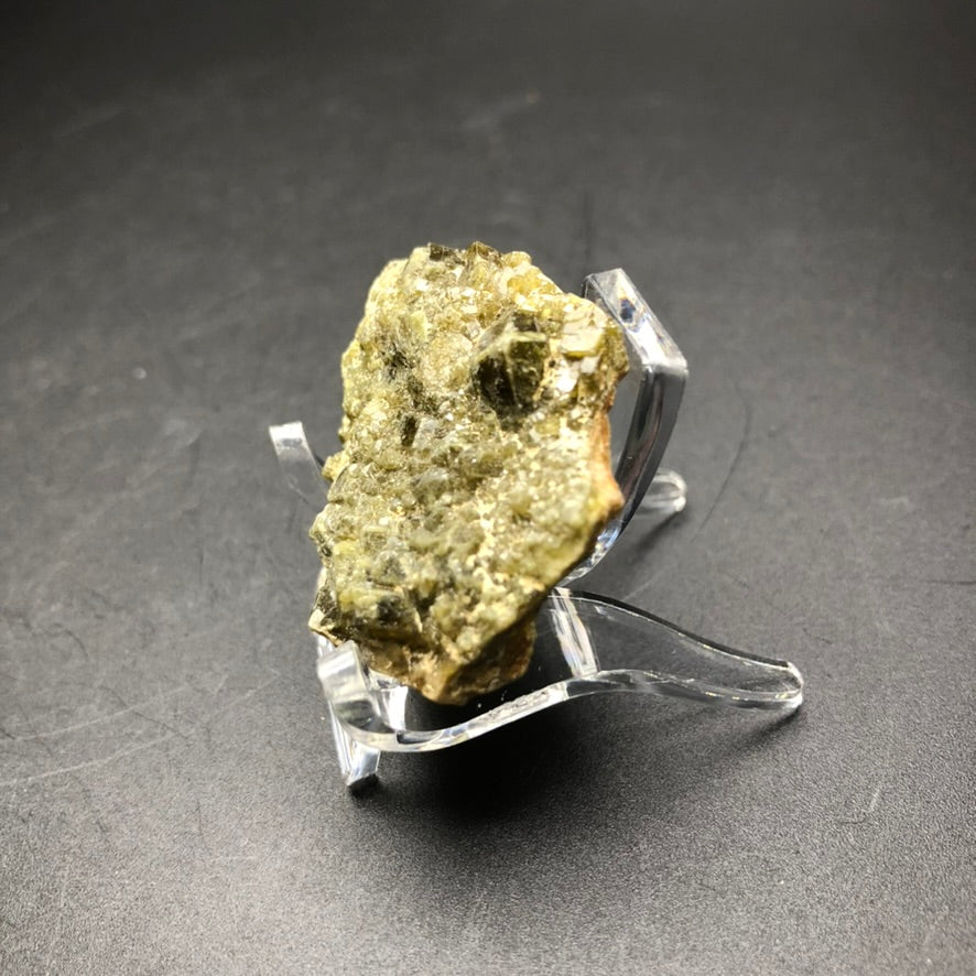 An uneven and rocky specimen, reminiscent of rare green garnets, sits on a transparent plastic stand against a dark gray background. This mineral, originating from Tubissis in the Erongo region of Namibia, is featured as an AUCTION item by The Crystalary: the rough Dermantoid Garnet.