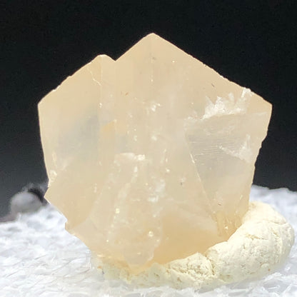 A light honey calcite crystal from Otero County, Colorado, by The Crystalary rests on a white surface. Its rough texture and translucent structure beautifully capture soft lighting against a dark background.