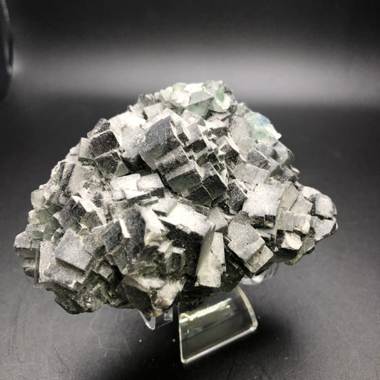 A cluster of dark gray and black mineral crystals, identified as the "AUCTION- Fluorite" from The Crystalary, sourced from the renowned Yaogangxian Mine in Yizhang County, Chenzhou, Hunan, China, showcases a cubic geometric pattern. It rests elegantly on a transparent stand against a plain dark background.