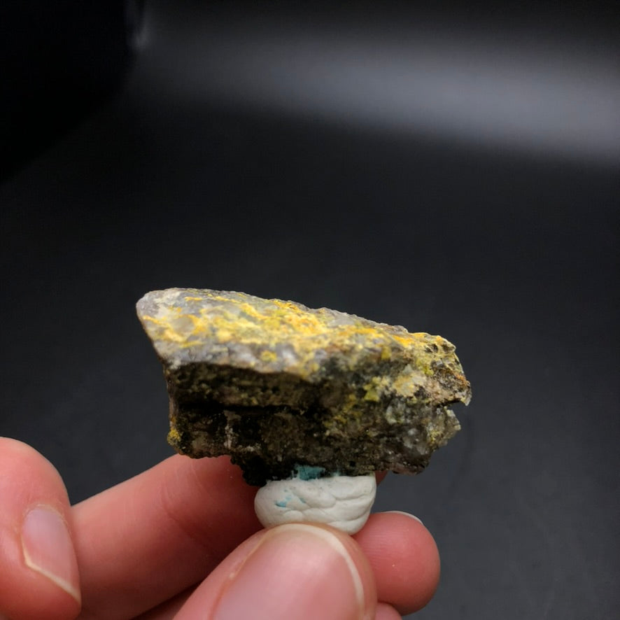 A close-up shot showcases a hand holding a rock specimen characterized by its rough texture and patches of yellow Pottsite minerals. The dark background accentuates the intricate details of the rock, which is said to have originated from the Linka Mine. This exquisite piece is featured in "AUCTION - Pottsite" offered by The Crystalary, sourced from Spencer Hot Springs Mining District, Lander County, Nevada, USA.