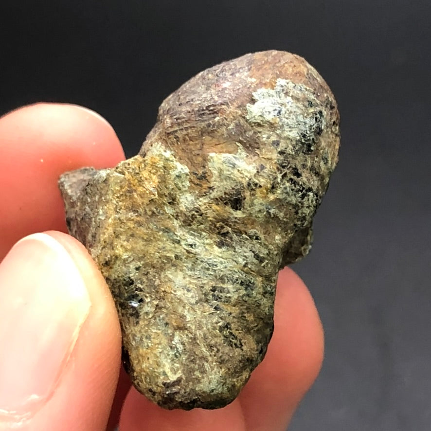 A hand holds The Crystalary's AUCTION-Almadine Garnet, featuring earthy shades of brown, beige, and green with garnet hues. Its textured surface stands out against a dark background, highlighting the natural beauty of this specimen from Markstay, Ontario.