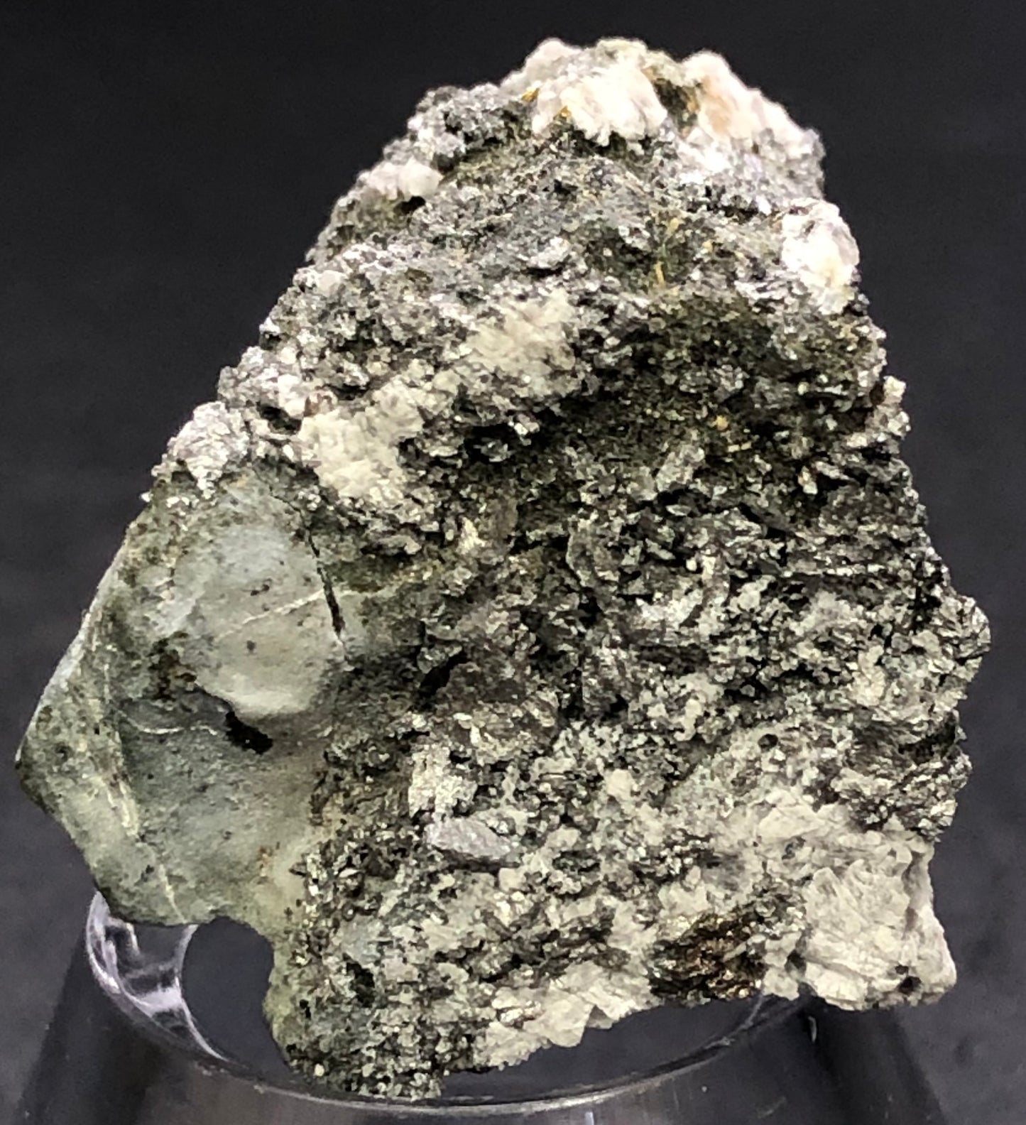 A close-up of a textured mineral specimen from The Crystalary, featuring gray and white hues, identified as pyrite or hematite. It rests on a small clear stand against a dark background, showcasing its raw allure. Identified as "AUCTION- Pyrite, hematite- unk locality.