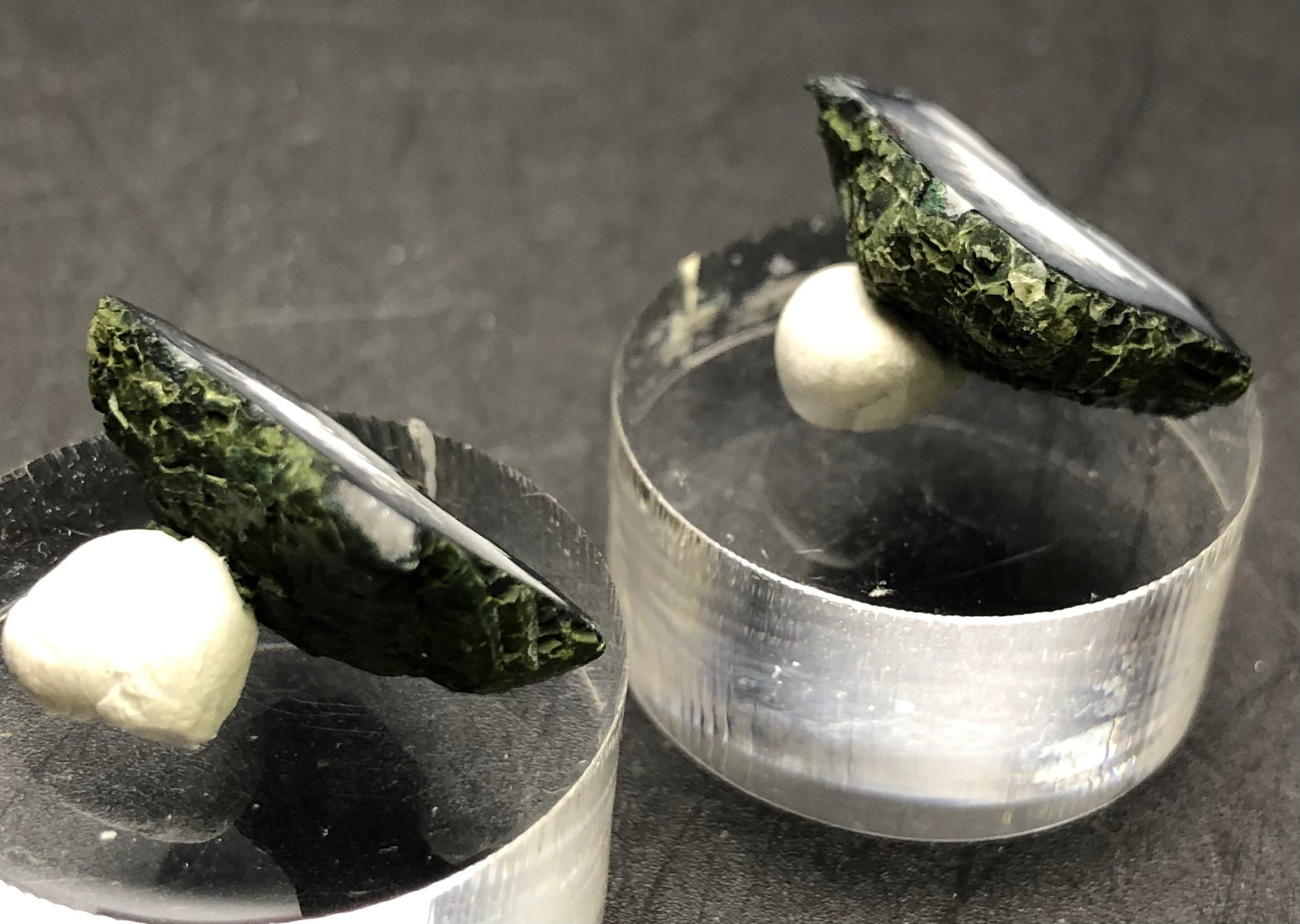 Two Tabasco geode samples from The Crystalary, characterized by their dark green texture similar to chalcedony, are elegantly mounted on small white supports and positioned on clear cylindrical stands. The backdrop features a simple, dark surface.