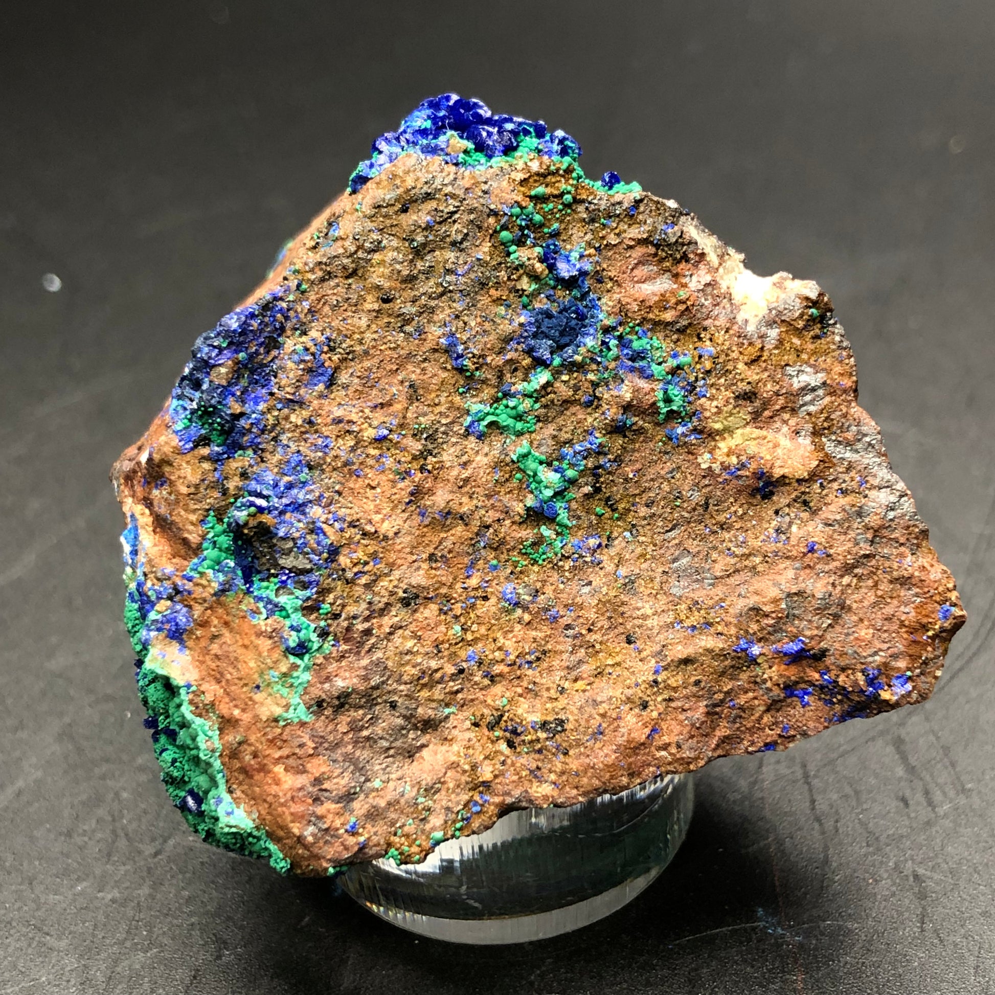 An impressive rock from The Crystalary's collection, showcasing vibrant azurite and malachite mineral deposits, rests on a small pedestal against a dark background. The minerals stand out in stark contrast to the rock's earthy texture, echoing treasures unearthed from the Apex Mine in Utah.