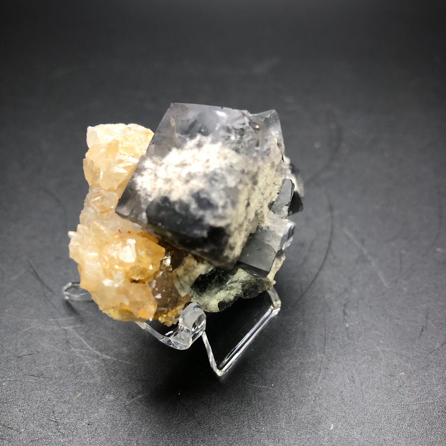 The Crystalary offers an item from the Milky Way Pocket at the Diana Marie Mine, featuring a striking, transparent cubic crystal with dark inclusions. This exquisite fluorite centerpiece boasts a smoky gray-blue hue and is surrounded by smaller yellowish and milky white crystals. Perfectly showcased against a dark backdrop, it rests on a clear stand.