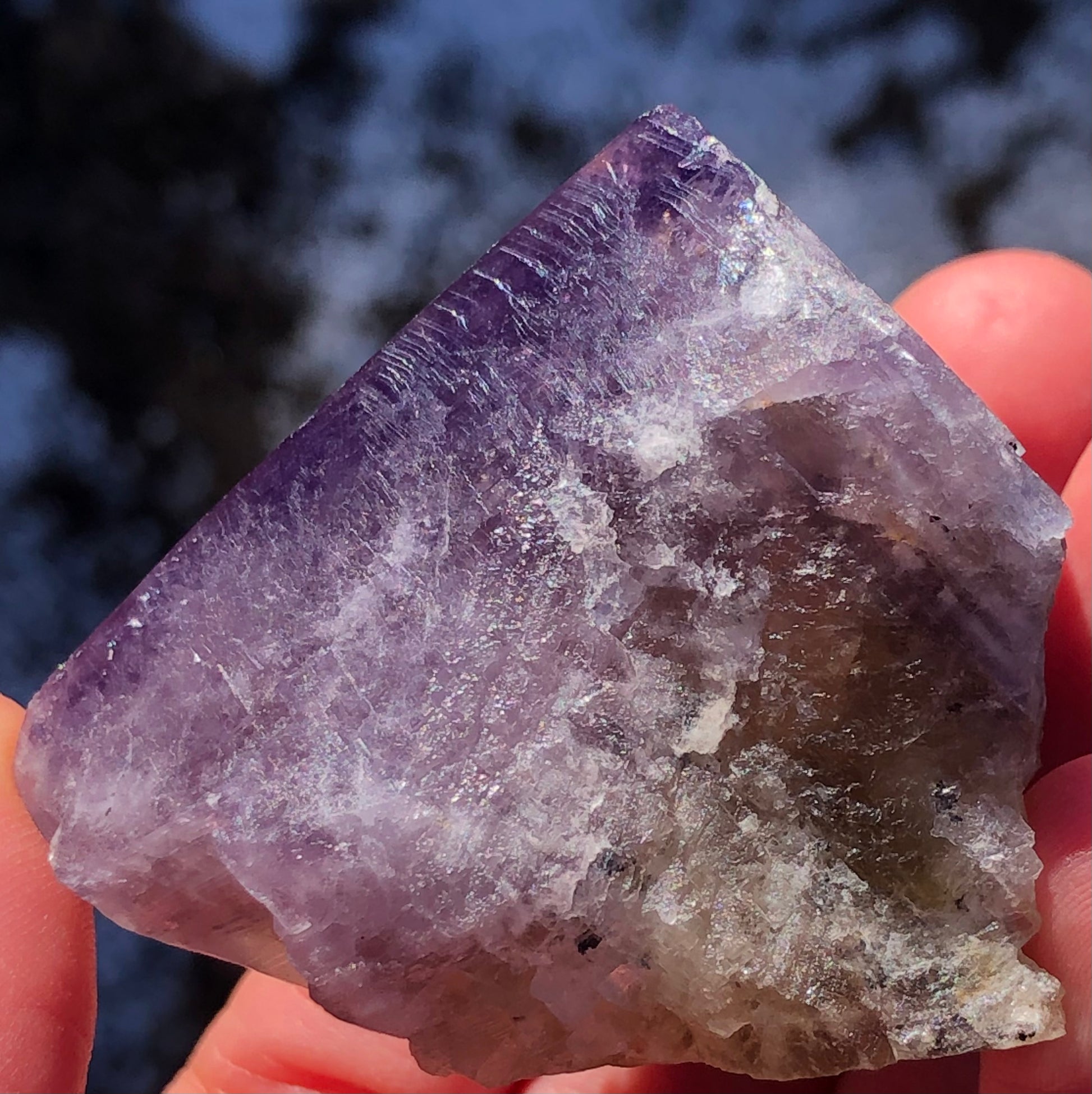 A hand displays a rough, triangular piece of The Crystalary's Fluorite from Cromwell Pocket, Greenlaws Mine. Against a blurred outdoor backdrop, the stone reveals multiple shades of purple with crystalline textures and a hint of translucency that brilliantly catches light.