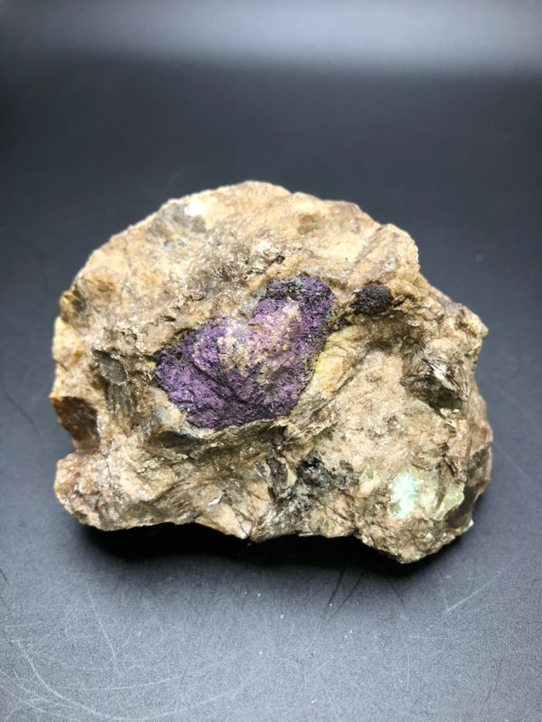 A rough brown rock featuring a prominent purple Wilsonite inclusion from "The Crystalary's AUCTION- Wilsonite- Canada," set against a plain dark background.