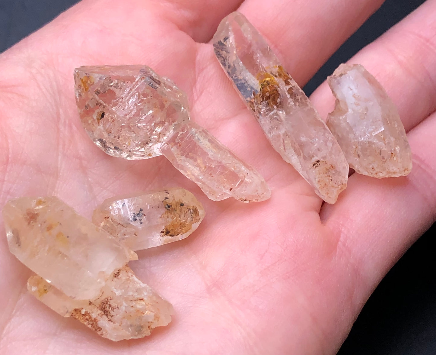 An AUCTION- Petroleum Quartz from The Crystalary, found in Afghanistan, is held by a hand. It features varying sizes with brown inclusions and rough surfaces that catch light elegantly, while a unique scepter-shaped piece glows distinctively under UV light.