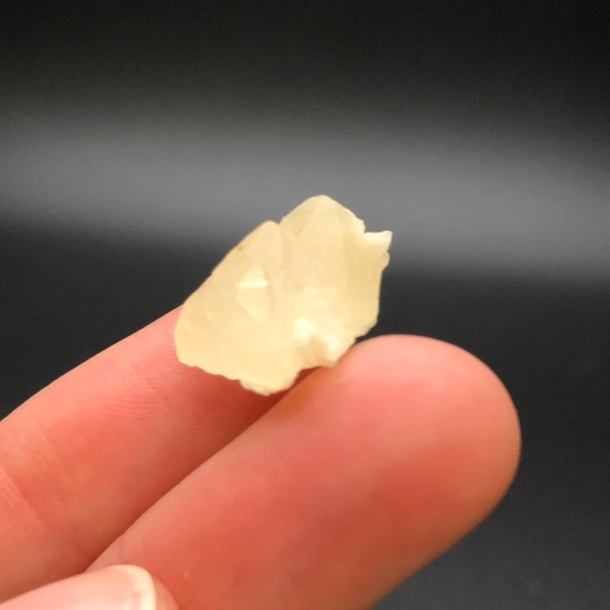 A person's fingers hold a small, opaque cream calcite crystal with a yellowish hue against a plain dark background. This unevenly shaped mineral, featuring rough edges, is sourced from the renowned Corydon Stone Co. Quarry in Indiana, USA and offered by The Crystalary under the product name "AUCTION- Calcite.