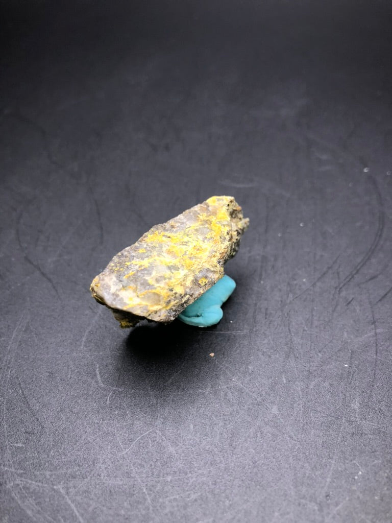 Displayed against a dark background, this rough stone features yellowish mineral deposits identified as Pottsite, set atop a small piece of blue clay. This piece originates from the Linka Mine and is part of "AUCTION- Pottsite- Linka Mine, Spencer Hot Springs Mining District, Lander County, Nevada, USA" by The Crystalary.