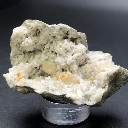 Capture the beauty of the AUCTION- Geode with Calcite from Worthington, Ohio, USA by The Crystalary. This stunning piece exhibits creamy white and pale green hues, featuring small, shiny orange and clear crystal formations akin to a druzy geode. It is elegantly displayed on a transparent stand against a dark background.