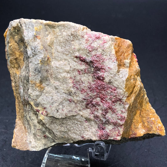 The Crystalary presents "AUCTION- Cinnabar- India," a rough, irregular rock with cinnabar deposits set against gray and brown stone, displayed on a clear stand with a black background to highlight its captivating matrix.