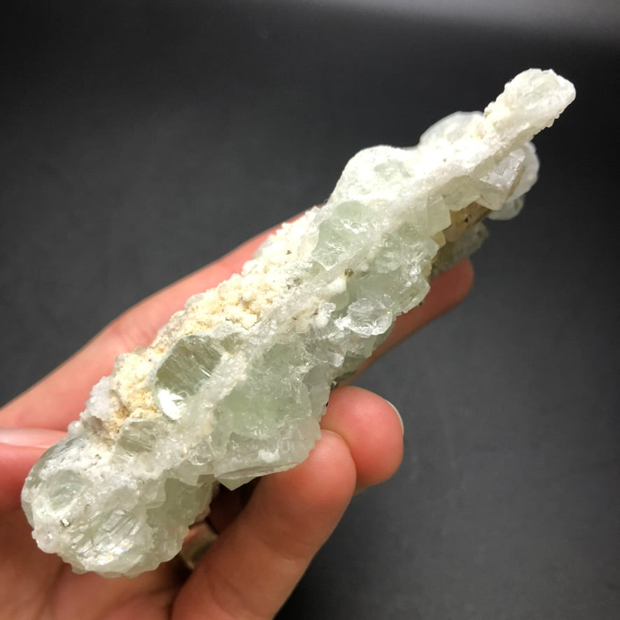 A hand holds The Crystalary's AUCTION- Fluorite mineral specimen against a dark background. This translucent, pale green and white specimen from the Xianghualing Mine in Linwu Co., Chenzhou, Hunan, China features clusters and jagged edges with crystalline textures that reveal hints of fluorite cubes nestled on a micro-quartz matrix.